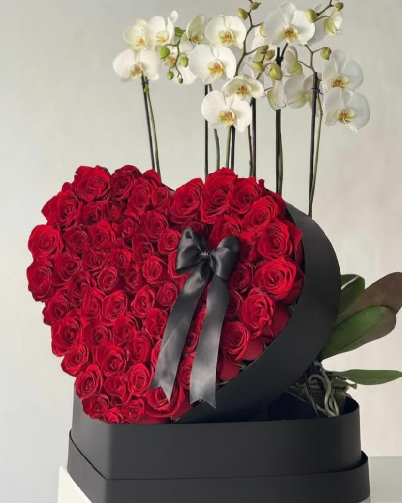 Heart-shaped arrangement of red roses with a black ribbon, surrounded by white orchids on a light gray background.